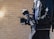 A person is holding a professional video camera with multiple attachments, including a monitor and stabilizer rig. The individual is wearing a black jacket and gloves, and has a backpack. The background consists of a light brick wall, suggesting an outdoor setting.