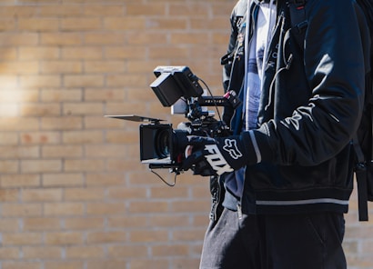 A person is holding a professional video camera with multiple attachments, including a monitor and stabilizer rig. The individual is wearing a black jacket and gloves, and has a backpack. The background consists of a light brick wall, suggesting an outdoor setting.