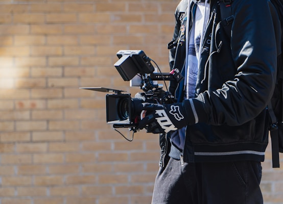 A person is holding a professional video camera with multiple attachments, including a monitor and stabilizer rig. The individual is wearing a black jacket and gloves, and has a backpack. The background consists of a light brick wall, suggesting an outdoor setting.