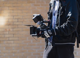 A person is holding a professional video camera with multiple attachments, including a monitor and stabilizer rig. The individual is wearing a black jacket and gloves, and has a backpack. The background consists of a light brick wall, suggesting an outdoor setting.