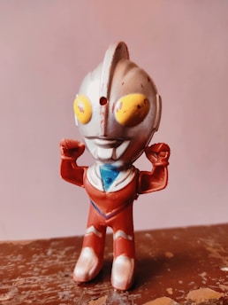 A small toy figure of a robot-like character with a metallic, humanoid appearance. It has a gray body with red and blue accents, large yellow eyes, and antennae-like structures on its head. The figure is in a standing pose with arms raised.