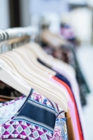 A collection of colorful clothes neatly arranged on wooden hangers displayed on a clothing rack. The garments feature various patterns and fabrics, showcasing a variety of styles.