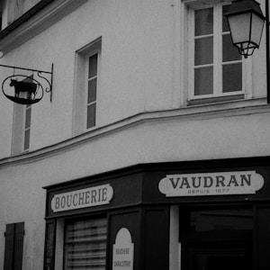 A vintage-style storefront with the word 'Boucherie' indicating a butcher shop. Above the shop, there is a sign with a silhouette of a cow. The architecture is classic with several windows and an old-fashioned lantern hanging on the side.