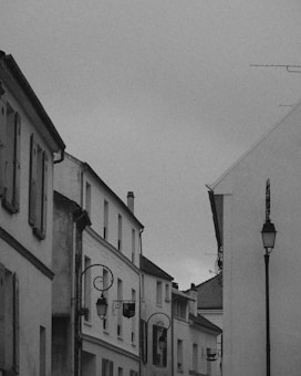 A black and white photograph of a narrow, quiet street lined with traditional European-style buildings. The architecture includes several old-fashioned street lamps with ornate designs, shuttered windows, and sloping rooftops. The sky above is overcast, contributing to the serene and somewhat nostalgic atmosphere.
