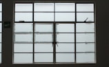 A large, rectangular window with multiple panes arranged in a grid pattern. The glass appears to be frosted or opaque, allowing minimal visibility to the outside. The window has simple metal latches for opening the panes, and there is a small green sign on the left side.
