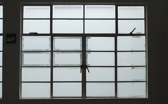 A large, rectangular window with multiple panes arranged in a grid pattern. The glass appears to be frosted or opaque, allowing minimal visibility to the outside. The window has simple metal latches for opening the panes, and there is a small green sign on the left side.