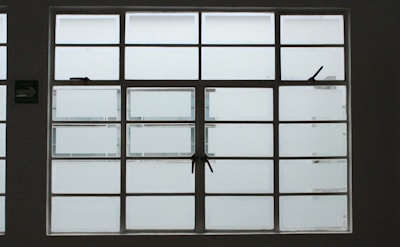 A large, rectangular window with multiple panes arranged in a grid pattern. The glass appears to be frosted or opaque, allowing minimal visibility to the outside. The window has simple metal latches for opening the panes, and there is a small green sign on the left side.