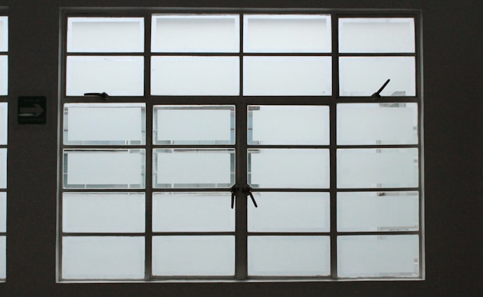 A large, rectangular window with multiple panes arranged in a grid pattern. The glass appears to be frosted or opaque, allowing minimal visibility to the outside. The window has simple metal latches for opening the panes, and there is a small green sign on the left side.