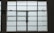 A large, rectangular window with multiple panes arranged in a grid pattern. The glass appears to be frosted or opaque, allowing minimal visibility to the outside. The window has simple metal latches for opening the panes, and there is a small green sign on the left side.