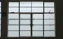 A large, rectangular window with multiple panes arranged in a grid pattern. The glass appears to be frosted or opaque, allowing minimal visibility to the outside. The window has simple metal latches for opening the panes, and there is a small green sign on the left side.