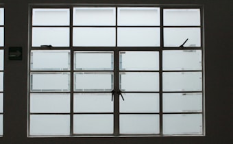 A large, rectangular window with multiple panes arranged in a grid pattern. The glass appears to be frosted or opaque, allowing minimal visibility to the outside. The window has simple metal latches for opening the panes, and there is a small green sign on the left side.