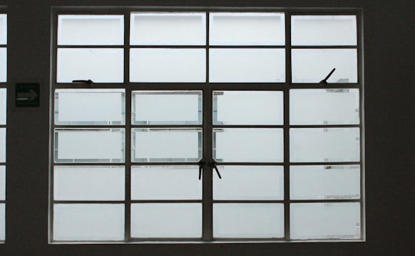 A large, rectangular window with multiple panes arranged in a grid pattern. The glass appears to be frosted or opaque, allowing minimal visibility to the outside. The window has simple metal latches for opening the panes, and there is a small green sign on the left side.