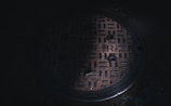 A circular metal sewer cover with a textured grid-like pattern and the word 'SEWER' embossed on its surface. The cover is situated on a dark, concrete ground. Subdued lighting casts a shadowy ambiance over the scene.