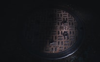 A circular metal sewer cover with a textured grid-like pattern and the word 'SEWER' embossed on its surface. The cover is situated on a dark, concrete ground. Subdued lighting casts a shadowy ambiance over the scene.