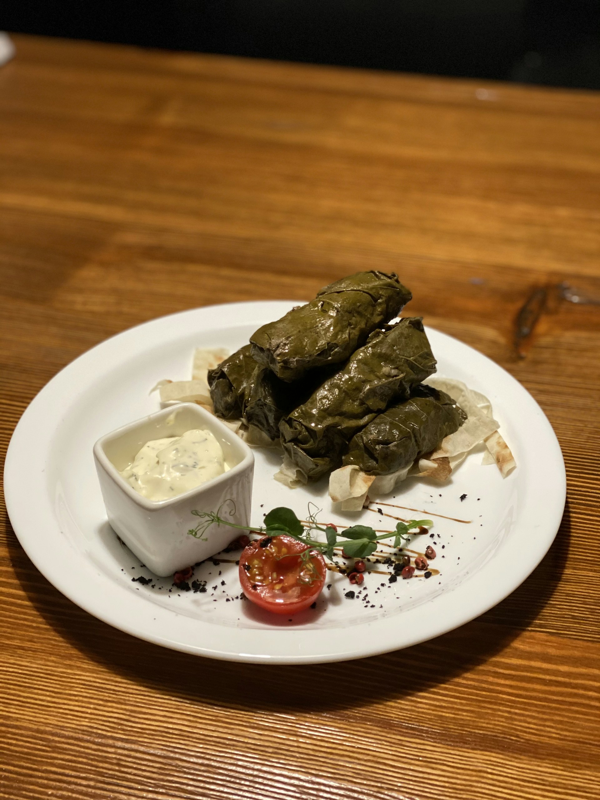 A white ceramic plate on a wooden table holds several grape leaf-wrapped rolls accompanied by a small white dish of creamy sauce. Garnish includes half a cherry tomato, microgreens, and a sprinkle of black peppercorns.