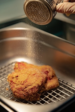 A gloved hand is using a shaker to sprinkle seasoning onto a piece of breaded meat resting on a metal rack inside a kitchen tray. The golden-brown crust of the meat contrasts with the metallic sheen of the shaker and tray, conveying a sense of careful preparation.