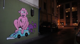 A piece of colorful street art on a building wall depicts a cartoon-like pink character joyfully riding a small blue race car. The character has an expressive face, while purple exhaust fumes trail from the vehicle. The urban setting is dimly lit, with parked cars and a large building in the background.
