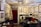 An artist's studio or cozy antique office filled with various framed paintings, photographs, and sketches on the walls. There is a desk with a computer, lamp, and scattered papers. Several chairs, including a yellow and red one, are placed around the room. The ceiling features an ornate light fixture, and the top of a red curtain can be seen on the back wall alongside art supplies and storage items.