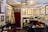 An artist's studio or cozy antique office filled with various framed paintings, photographs, and sketches on the walls. There is a desk with a computer, lamp, and scattered papers. Several chairs, including a yellow and red one, are placed around the room. The ceiling features an ornate light fixture, and the top of a red curtain can be seen on the back wall alongside art supplies and storage items.