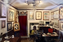 An artist's studio or cozy antique office filled with various framed paintings, photographs, and sketches on the walls. There is a desk with a computer, lamp, and scattered papers. Several chairs, including a yellow and red one, are placed around the room. The ceiling features an ornate light fixture, and the top of a red curtain can be seen on the back wall alongside art supplies and storage items.