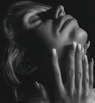 A close-up black-and-white photograph of a person's face and hand. The person holds their hand gracefully against their chin, with fingers extended and adorned with manicured fingernails that have a subtle design. The lighting casts soft shadows, emphasizing the texture of the skin and the delicate details of the nails.