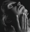 A close-up black-and-white photograph of a person's face and hand. The person holds their hand gracefully against their chin, with fingers extended and adorned with manicured fingernails that have a subtle design. The lighting casts soft shadows, emphasizing the texture of the skin and the delicate details of the nails.