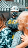 A person is wearing a protective helmet with a face mask and a uniform that includes a yellow vest. They are standing among a crowd, with a striped canopy in the background.