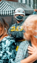 A person is wearing a protective helmet with a face mask and a uniform that includes a yellow vest. They are standing among a crowd, with a striped canopy in the background.
