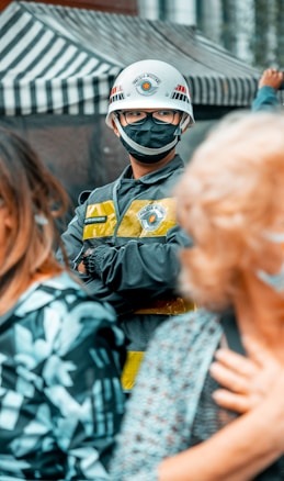 A person is wearing a protective helmet with a face mask and a uniform that includes a yellow vest. They are standing among a crowd, with a striped canopy in the background.