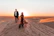 A couple walking hand-in-hand across golden desert dunes at sunset.