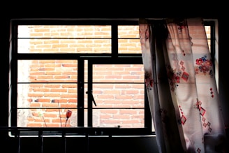 A large window with black framing is partially covered by a floral-patterned curtain. Behind the window, a brick wall is visible, partially sunlit and casting shadows. Some small objects can be seen in silhouette against the bottom edge of the window.