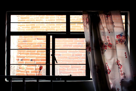 A large window with black framing is partially covered by a floral-patterned curtain. Behind the window, a brick wall is visible, partially sunlit and casting shadows. Some small objects can be seen in silhouette against the bottom edge of the window.