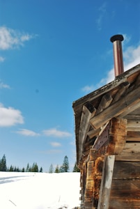 A rustic log cabin with a metal chimney set against a bright blue sky. The cabin is partially covered by snow, and evergreen trees are visible in the background. The logs are weathered, giving the cabin a vintage feel.