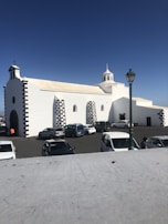 A white church with black stone accents is situated in a parking lot with several parked cars and a green street lamp. The building features multiple arched windows and a small, central bell tower set against a clear blue sky.