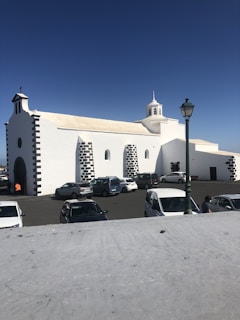 A white church with black stone accents is situated in a parking lot with several parked cars and a green street lamp. The building features multiple arched windows and a small, central bell tower set against a clear blue sky.