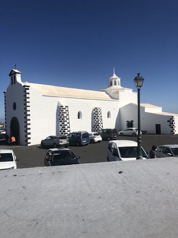 A white church with black stone accents is situated in a parking lot with several parked cars and a green street lamp. The building features multiple arched windows and a small, central bell tower set against a clear blue sky.