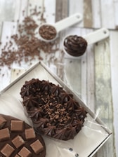 A variety of chocolate desserts are artfully arranged. There are two main items, one topped with smooth swirls and sprinkled with chocolate shavings and the other decorated with chocolate squares. In the background, there's chocolate sprinkles spread out on the wooden surface and a measuring spoon filled with more chocolate sprinkles.