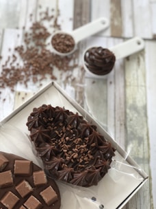 A variety of chocolate desserts are artfully arranged. There are two main items, one topped with smooth swirls and sprinkled with chocolate shavings and the other decorated with chocolate squares. In the background, there's chocolate sprinkles spread out on the wooden surface and a measuring spoon filled with more chocolate sprinkles.