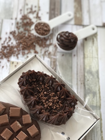 A variety of chocolate desserts are artfully arranged. There are two main items, one topped with smooth swirls and sprinkled with chocolate shavings and the other decorated with chocolate squares. In the background, there's chocolate sprinkles spread out on the wooden surface and a measuring spoon filled with more chocolate sprinkles.