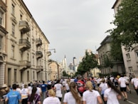 A large group of people are running on a street flanked by residential buildings. The runners are wearing athletic clothing and race bibs, indicating that this is likely an organized event. The cityscape can be seen in the background, with tall buildings visible on the skyline. The sky is overcast, creating a subdued lighting.