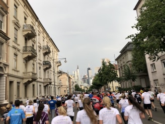 A large group of people are running on a street flanked by residential buildings. The runners are wearing athletic clothing and race bibs, indicating that this is likely an organized event. The cityscape can be seen in the background, with tall buildings visible on the skyline. The sky is overcast, creating a subdued lighting.