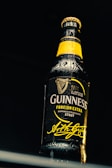 A detailed close-up of a dark glass beer bottle under dramatic lighting. The bottle features a prominent yellow label and highlights the brand name with a signature beneath.