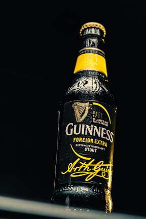 A detailed close-up of a dark glass beer bottle under dramatic lighting. The bottle features a prominent yellow label and highlights the brand name with a signature beneath.
