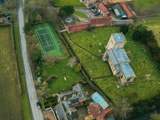 An aerial view of a countryside area featuring an old stone church with a graveyard around it. Adjacent to the church is a residential building with a fenced garden. A tennis court with a vivid green surface is situated nearby. Several parked cars are visible along with trees and open grassy areas. The scene is bordered by a road at the edge.