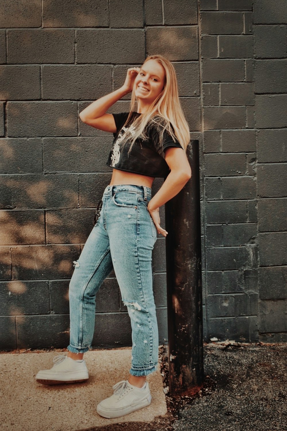 woman in black tank top and blue denim jeans leaning on brown brick wall