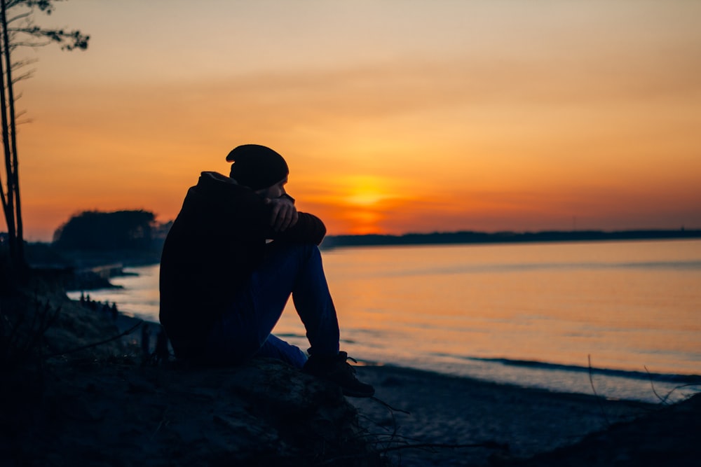 silhouette of man sitting on rock near body of water during sunset