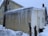 A small house with light-colored siding is covered with long icicles hanging from the roof. Snow surrounds the house, indicating a cold and wintry setting. There is a brick chimney and a satellite dish attached to the side of the house.