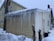 A small house with light-colored siding is covered with long icicles hanging from the roof. Snow surrounds the house, indicating a cold and wintry setting. There is a brick chimney and a satellite dish attached to the side of the house.