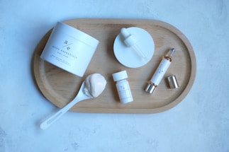 A neatly arranged skincare set featuring various products on a wooden tray. Included are a jar of body cream, a dropper bottle, a small bottle, a spoon with cream, and a cylindrical bottle with a silver cap. The background is a light gray textured surface.