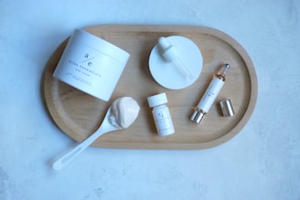 A neatly arranged skincare set featuring various products on a wooden tray. Included are a jar of body cream, a dropper bottle, a small bottle, a spoon with cream, and a cylindrical bottle with a silver cap. The background is a light gray textured surface.
